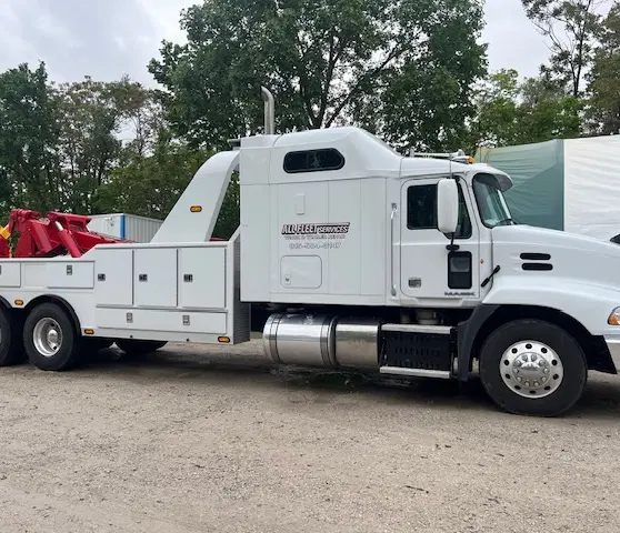 A white tow truck with a red tow arm parked on a gravel surface, surrounded by trees.